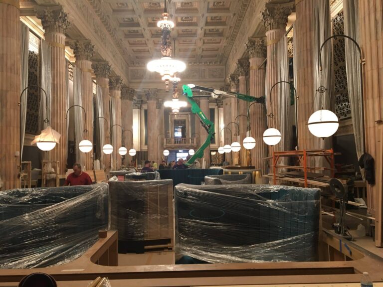 Interior of a historic building under construction with tall ornate columns, chandeliers, and protective plastic covering furniture. Workers and construction equipment are visible, with rows of hanging spherical lights illuminating the space.