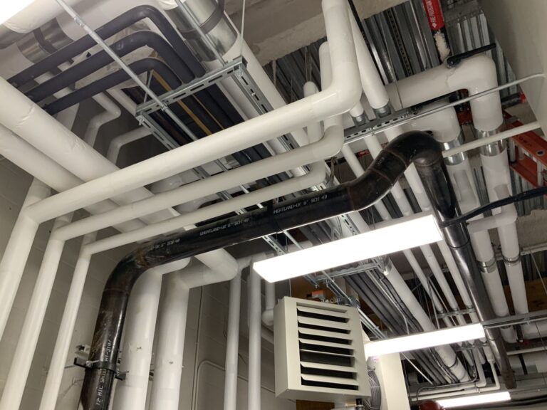 An industrial ceiling with a complex network of white and black pipes and ducts. Fluorescent lighting fixtures illuminate the area, highlighting metal and plastic components. A ventilation unit is also visible among the network of pipes.