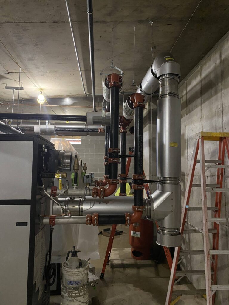 A complex network of newly installed industrial pipes in a utility room, featuring a mix of black and silver piping with red connectors. A concrete ceiling with exposed lighting is visible above, alongside construction equipment such as a ladder and bucket in the foreground, highlighting an active construction or maintenance setting.
