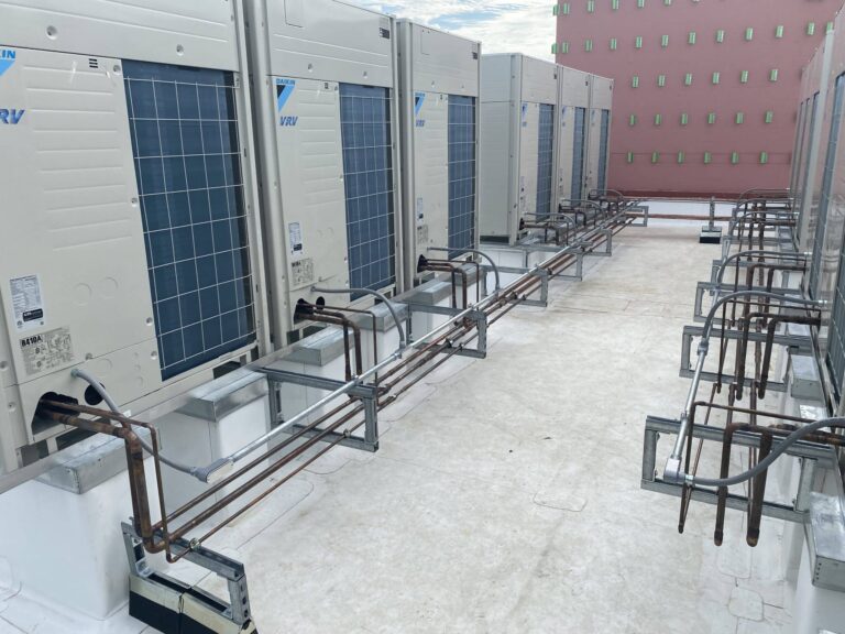 A row of large commercial air conditioning units installed on a rooftop, each equipped with multiple visible pipes and connectors, set against a clear blue sky and adjacent to a building with a pink facade marked by small, green-bordered windows.